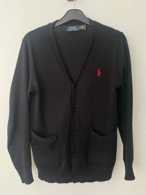 Svart cardigan från Polo Ralph Lauren - Säljer nu en riktigt snygg klassisk svart cardigan från Ralph Lauren, då den tyvärr har blivit för liten. Tröjan har två fickor framtill, ribbade muddar och den klassiska röda logotypen broderad på bröstet. Storleken är M men liten i storleken, passar runt 180. Den är använd men fortfarande i gott skick, finns en liten fläck på armen men inget som syns när man har på den. Skriv för frågor om mått, pris eller fler bilder! 