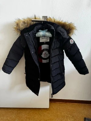 Svart dunjacka med fuskpäls - Snygg svart dunjacka  med avtagbar huva och fuskpäls. Jackan har dragkedja och knappar framtill samt quiltad design. Perfekt för kalla dagar och riktigt varm.