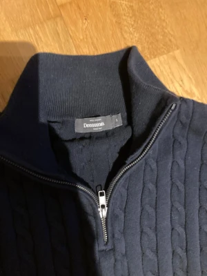 Mörkblå kabelstickad tröja Dressmann - Mörkblå kabelstickad tröja från Dressmann med halv dragkedja och hög krage. Tröjan har långa ärmar och ribbade muddar vid ärmslut och nederkant. Perfekt för dig som gillar klassisk och stilren look.