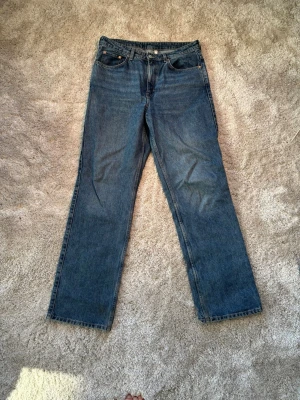 Blå raka jeans i klassisk modell - Säljer ett par blå jeans med rak passform och fem fickor. Jeansen har en klassisk look med normal midja och knappgylf. Hög midja i modellen ROWE från Weekday i extra lång modell (34)