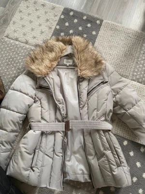 Ljusgrå pufferjacka med fuskpäls - Snygg ljusgrå pufferjacka från Isolde med avtagbar fuskpäls på kragen och bälte i midjan för en markerad siluett. Jackan har två bröstfickor med dragkedja och quiltad design som ger extra värme. Perfekt för kalla vinterdagar. XL-L