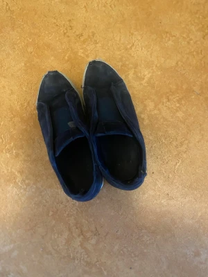 Mörkblå sneakers från Arigato - Säljer ett par mörkblå sneakers från Arigato med vit sula och klassisk slip-on design. Skorna har ovandel i tyg och elastiska band över vristen för enkel påtagning. Perfekta för en avslappnad streetstil.