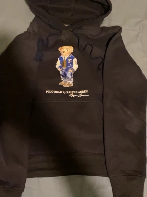 Mörkblå Polo Bear hoodie Ralph Lauren - Mörkblå hoodie från Polo Ralph Lauren med den ikoniska Polo Bear-trycket på bröstet. Hoodien har dragsko i huvan och en stor magficka. Perfekt för dig som vill ha en klassisk streetwear-look med en twist.
