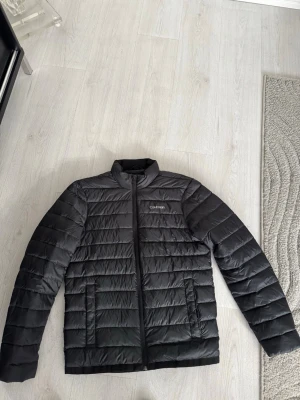 Svart dunjacka från Calvin Klein - Snygg svart dunjacka från Calvin Klein med diskret logga på bröstet. Jackan har ståkrage, dragkedja framtill och två stora fickor. Quiltad design med horisontella sömmar ger en modern look. Perfekt för kalla vinterdagar.