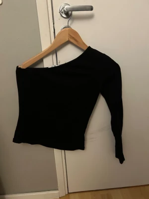 Svart oneshoulder topp från NA-KD - Snygg svart oneshoulder topp från NA-KD. Toppen har lång ärm på ena sidan och är axelbandslös på andra💞