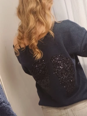 Mörkblå stickad tröja med paljetter vingar  -  SÖKER!!mörkblå stickad tröja med off shoulder-design och långa ärmar. På ryggen finns ett stort paljettmönster i form vingar 