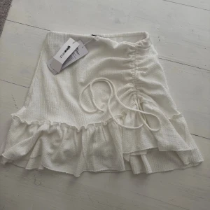 Vit volangkjol med snörning från Gina Tricot - Supersöt vit kortkjol från Gina Tricot med volangkant och snörning i sidan. Kjolen har en lätt struktur i tyget och är perfekt för sommarens alla outfits. Den är helt ny med lappen kvar och säljs då den var för liten.