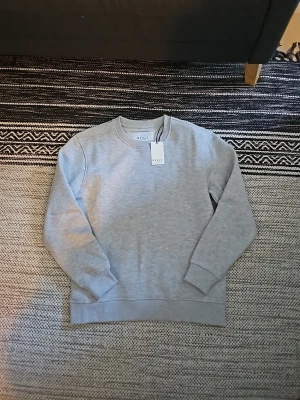 Ljusgrå sweatshirt från RYVLS - En stilren ljusgrå sweatshirt från RYVLS Stockholm. Tröjan har rund halsringning, ribbade muddar vid ärmslut och nederkant samt en mjuk bomullskänsla. Perfekt för en clean och avslappnad look. Inga tryck eller mönster, bara enkel och snygg design.