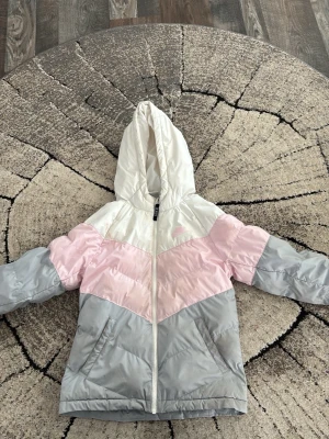 Pastellfärgad dunjacka med huva - Snygg dunjacka i pastellfärger med huva och dragkedja framtill. Jackan har blockmönster i vitt, ljusrosa och ljusgrått samt två sidofickor. Perfekt för kalla vinterdagar och ger en trendig look.