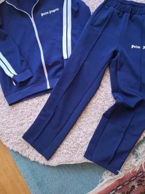 Blå tracksuit från Palm Angels - Säljer ett par blåa tracksuits från Palm Angels med vit logga på ena benet. Byxorna har resår i midjan och raka ben. Materialet är mjukt och stretchigt. Klassisk sportig design med enkel stil.