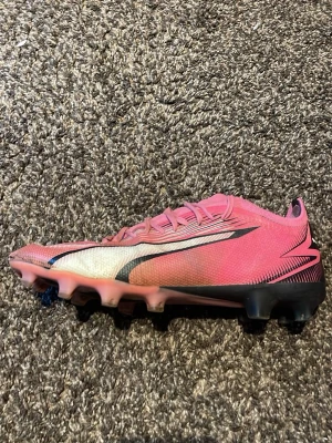 Rosa Puma fotbollsskor med dobbar - Säljer ett par snygga rosa fotbollsskor från Puma med svart och vit detalj på sidan. Skorna har låg profil, snörning och slitstark ovandel i syntetmaterial. Dobbarna är utformade för bra grepp på gräsplan. Perfekta för dig som vill sticka ut på planen!