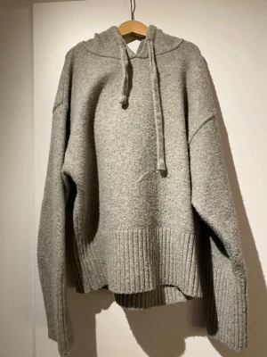 Grå stickad hoodie med luva - Mysig grå stickad hoodie med luva och dragsko. Tröjan har breda ribbstickade muddar vid ärmslut och nederkant samt avslappnad passform. Perfekt för kalla dagar när du vill vara både bekväm och snygg.