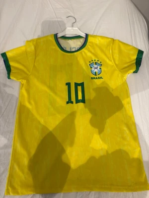 Brasilien tröja Pelé nr 10 gul - Gul Brasilien fotbollströja med gröna detaljer, nummer 10 och Pelé på ryggen. Tröjan har CBF-logga på bröstet och klassisk rund hals. Perfekt för dig som älskar fotboll och vill hylla en legend. Tillverkad i lätt och bekvämt material.