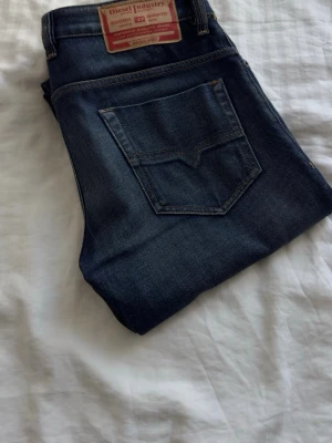 Mörkblå jeans från Diesel - Snygga mörkblå jeans från Diesel med klassisk femficksmodell och diskreta slitningar framtill. Jeansen har raka ben och är tillverkade i ett denimtyg som ger en bekväm passform. Perfekta för dig som gillar en tidlös och stilren look.