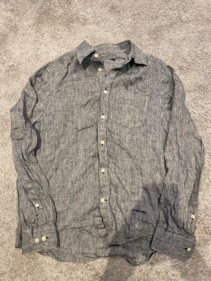 Grå linneskjorta från J.Lindeberg - Snygg grå skjorta från J.Lindeberg i svalt linne. Klassisk krage, vita knappar och diskret logga broderad nedtill. Perfekt till jeans eller chinos för en clean och avslappnad look.