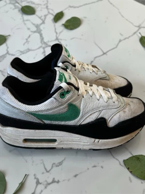 Nike Air Max 1 vita och gröna sneakers - Nike Air Max 1 sneakers i vitt och svart med gröna detaljer och klassisk grön Swoosh. Ovandelen är i mesh och mocka, med vita skosnören och svart sula. Ikonisk retrostil som passar perfekt till streetwear och casual outfits.