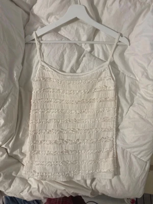 Vit smockad linnetopp med tunna band - Säljer en söt vit linnetopp med smock och tunna axelband. Toppen har ett diskret strukturmönster och är perfekt för varma dagar. Passar till jeans, kjol eller shorts för en fräsch look.