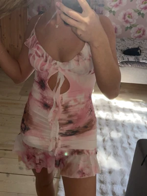 Rosa tie-dye klänning med volanger - Söt klänning i rosa och vitt tie-dye-mönster med tunna axelband, volangkant och dekorativ knytning framtill. Djup urringning och liten cut-out detalj under bysten. Kort modell med volang nertill, perfekt för mysiga kvällar.