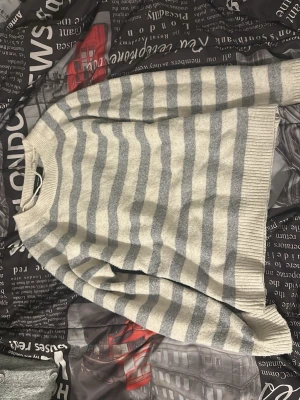 Randig stickad tröja i grått och vitt - Mysig stickad tröja med breda horisontella ränder i ljusgrått och vitt. Tröjan har rund halsringning och ribbade muddar vid ärmslut och nederkant. Perfekt för lager på lager och chill dagar.