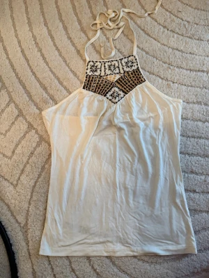 Vit halterneck topp med brodyr - Säljer en vit halterneck topp med snygg broderad detalj i svart och beige vid halsen. Toppen har tunna knytband i nacken och är tillverkad i ett mjukt, lätt tyg som känns skönt mot huden. Perfekt för varma dagar och en avslappnad stil.