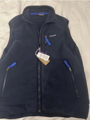 Mörkblå fleeceväst från Patagonia - Snygg mörkblå fleeceväst från Patagonia med dragkedja framtill och tre fickor med blå detaljer på dragkedjorna. Västen har hög krage och är tillverkad i mjukt, värmande fleecematerial. Perfekt för lager-på-lager och outdoor.