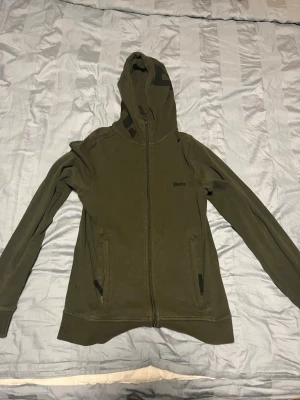 Olivgrön hoodie från Bench - Snygg olivgrön hoodie från Bench med dragkedja framtill och två fickor med svarta detaljer. Hoodien har en klassisk huva och diskret Bench-logga på bröstet. Perfekt för en avslappnad och cool stil.