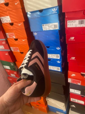 Hummel VTG CPH Sneakers Svart/Rosa EU 42 - Snygga svarta sneakers från Hummel med klassiska rosa chevrons på sidan och brun gummisula. Skon har vita skosnören och en låg profil som ger en retro känsla. Materialet är en mix av mocka och textil för en cool och sportig look.