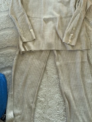 Beige ribbat loungewear-set - Mysigt loungewear-set i beige med tunika och byxor. Setet har smala ribbade ränder och knappar vid ärmsluten på toppen. Perfekt för chill dagar hemma. Mjukt och stretchigt material för extra komfort.