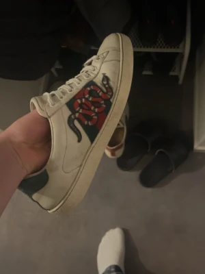 Gucci Ace sneakers - Snygga Gucci Ace sneakers i vitt läder med den ikoniska gröna och röda sidoremsan och en broderad orm. Skon har vita snören, låg profil och detaljer i grönt och rött på hälen. Perfekt för dig som vill sticka ut med lyxig streetstyle. Storlek 41. Lite av lädret har skaft men man tänker inte på de när de sitter på.