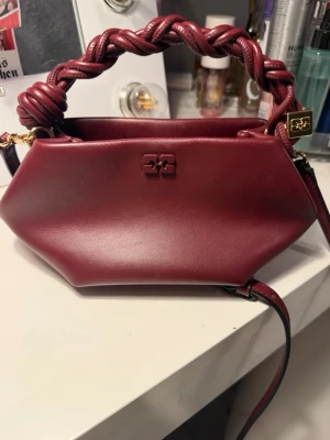 Vinröd handväska från Ganni  - Snygg vinröd handväska från Ganni. Väskan har ett flätat handtag och guldfärgade detaljer, inklusive logotypen framtill. Perfekt accessoar för att lyfta din outfit.