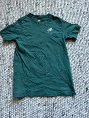 Grön Nike t-shirt - Snygg grön t-shirt från Nike med klassisk logga på bröstet. T-shirten har rund halsringning och korta ärmar. Tillverkad i mjuk bomull som är skön att bära. Perfekt till jeans eller shorts för en avslappnad stil.