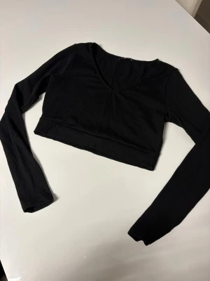 Svart croppad långärmad topp - Snygg svart croppad topp med lång ärm och v-ringning. Tillverkad i ribbad bomullsmix som ger en skön och stretchig passform. Perfekt att styla med höga jeans eller kjol för en trendig look.