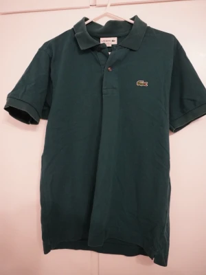 Mörkgrön pikétröja från Lacoste - Lacoste mörkgrön piké Size US XS  Säljer en riktigt fin Lacoste piké i mörkgrön färg. Klassisk modell med bra passform, funkar till både vardag och mer uppklätt. Använd lite men fortfarande i väldigt bra skick.  Sjukt clean färg och sitter riktigt snyggt på. Perfekt nu till vår/sommar.  Storlek: US XS Skick: 8/10 Pris: (100)