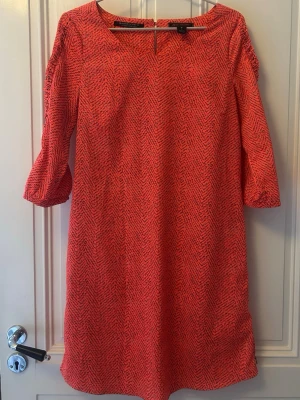 Röd/orange mönstrad klänning från Maison Scotch - Säljer en snygg orange klänning från Maison Scotch med diskret mönster och trekvartsärmar. Klänningen har en rak passform och rund halsringning, fin liten volangdetaljer sydd på ärmen. Perfekt för dig som gillar stilrena och färgstarka plagg. Materialet känns mjukt och lätt mot huden. 100% Polyester