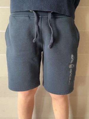 Mörkblå shorts från Sail Racing - Mörkblå shorts från Sail Racing med snörning i midjan och tryckt logga på ena benet. Shortsen har en bakficka och är tillverkade i mjuk bomullsmix som passar perfekt för sommardagar. Klassisk och avslappnad passform. OBS! Barnstorlek 150
