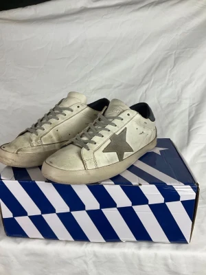 Vita Golden Goose sneakers med stjärna - Snygga vita sneakers från Golden Goose med grå stjärna på sidan och grå skosnören. Skorna har en klassisk låg profil och svart detalj vid hälen. Tillverkade i läder med slitna detaljer för en trendig look.