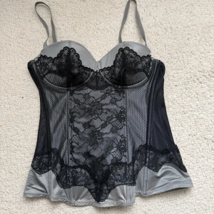 Silvergrå korsettopp med spets - Snygg silvergrå / svart korsettopp med svarta spetsdetaljer och tunna axelband. Toppen har genomskinliga meshpartier på sidorna och vacker spets framtill och längs bysten. Perfekt för dig som vill ha en edgy och elegant look.