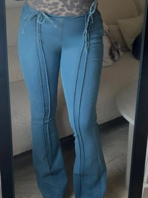 Blå utsvängda jeans med snörning - Säljer ett par ljusblå jeans med utsvängda ben och dekorativa snörningar i midjan. Jeansen har markerade sömmar framtill som ger en cool retrovibe. Perfekta för dig som gillar en unik och trendig stil. Endast provade då jag tyckte de var lite för små för mig. 