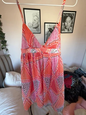 Färgglad kort sommarklänning - Säljer en kort klänning med smala axelband och v-ringning. Klänningen har ett färgstarkt mönster i rosa, orange och vitt med inslag av lila. Materialet känns lätt och luftigt, perfekt för varma dagar. Passformen är lös och avslappnad. Från H&m och i stl S!