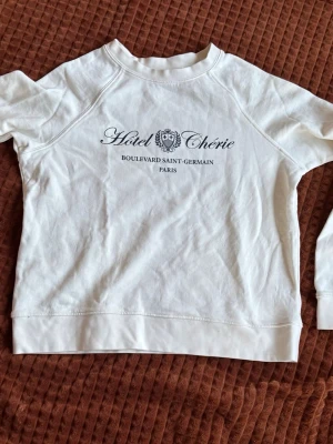 Vit sweatshirt med tryck Hotel Chérie - Vit sweatshirt med svart texttryck 'Hotel Chérie Boulevard Saint-Germain Paris' på bröstet. Tröjan har rund halsringning, långa ärmar och ribbade muddar. Tillverkad i mjukt bomullsmaterial för en avslappnad look.