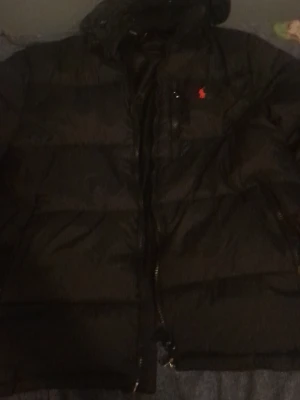 Svart dunjacka från Polo Ralph Lauren - Snygg svart dunjacka från Polo Ralph Lauren med klassisk röd logga på bröstet. Jackan har huva, dragkedja framtill och quiltad design som håller värmen. Perfekt för kalla dagar och riktigt stilren look. Inge skada elle nått sånt använd en gång men passade inte mig