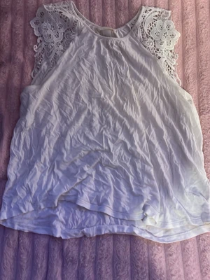 Vit ärmlös topp med spetsdetaljer - Säljer en vit ärmlös topp med vackra spetsdetaljer på axlarna. Toppen har en lös passform och rund halsringning. Perfekt för dig som gillar en feminin och luftig stil.