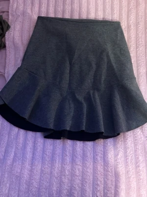 Mörkgrå volangkjol med dragkedja - Supersnygg mörkgrå kjol med volangkant och figurnära passform. Kjolen har en synlig dragkedja bak och är gjord i ett stretchigt, mjukt material som ger en bekväm känsla. Perfekt för dig som gillar en trendig och stilren look.