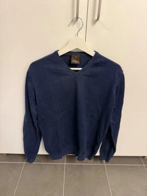 Blå V-ringad tröja Oscar Jacobson, stl M - Mörkblå stickad tröja från Oscar Jacobson med V-ringning och långa ärmar. Tröjan har ribbade muddar vid hals, ärmslut och nederkant. Perfekt för dig som gillar stilrena och klassiska plagg.