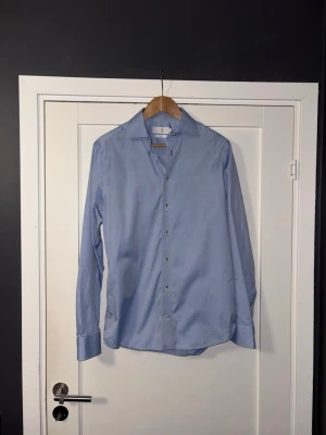 Ljusblå skjorta från Riley Slim Fit - Snygg ljusblå skjorta från Riley i slim fit-modell. Skjortan har klassisk krage, långa ärmar och knappar framtill. Perfekt för dig som gillar stilrena och enkla plagg. Tillverkad i mjuk bomull för skön känsla.