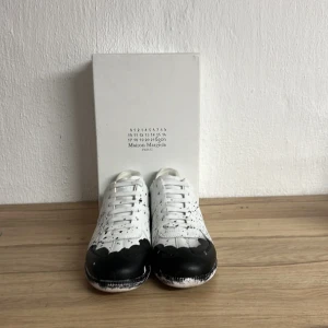Maison Margiela svartvita sneakers - Unika sneakers från Maison Margiela i vitt läder med svart färgstänk och svartmålade tår. Klassisk låg modell med vita skosnören och subtila detaljer som ger en konstnärlig vibe. Perfekta för dig som vill sticka ut med din stil. Otroligt bra skick på dem och använda 2 gånger. Perfekta nu till sommaren! Skicka vid minsta fundering. Mvh Lion 