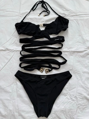 Svart cut-out baddräkt Dolce & Gabbana - Snygg svart baddräkt från Dolce & Gabbana med flera cut-out detaljer och långa band som snöras runt midjan. Baddräkten har guldfärgade DG-detaljer och halterneck knytning. Tillverkad i stretchigt material som passar perfekt för stranden eller poolen.