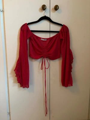Röd topp med volangärm - Röd topp från ZARA med långa volangärmar och rynkdetalj framtill med knytband. Tillverkad i ett lätt, transparent tyg som ger en luftig känsla. Aldrig använd.