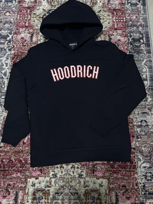 Svart HOODRICH hoodie med tryck - Svart hoodie från Hoodrich med stor vit och röd logga broderad på bröstet. Klassisk huva och magficka framtill. Perfekt för dig som gillar streetwear och vill ha en stilren men ändå iögonfallande look.