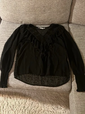 Svart blus med volangdetaljer - Snygg svart blus med långa ärmar och volangdetaljer längs V-ringningen. Blusen är tillverkad i ett tunt, transparent tyg och har smockade muddar vid ärmsluten. Använde  ett Linne under då den är lite genomskinlig!💕